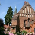 Backsteinkirche mit spitzem Turm und prächtigen Rosengärten, eingerahmt von blauem Himmel und Bäumen.