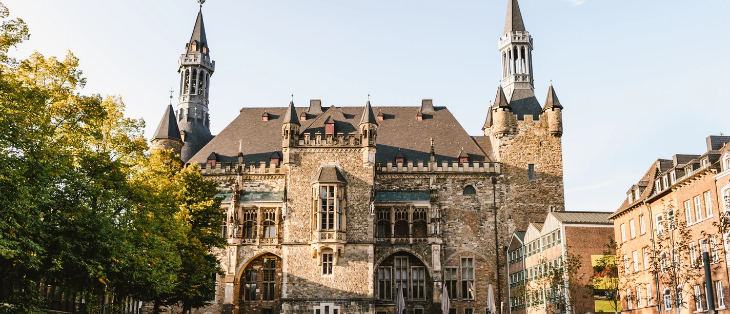 Aachener Rathaus auf Katschhof mit prächtiger Fassade aus Bruchstein, umgeben von alten Bäumen und historischer Architektur.