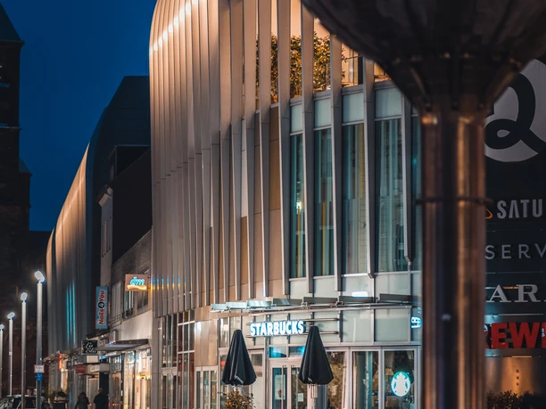 Nachts erleuchtetes Einkaufszentrum "Aquis Plaza" mit modernen Fassadenelementen und Geschäften.