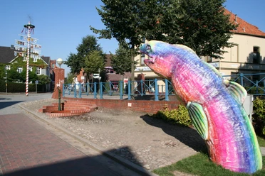 Bunte Skulptur eines Aals auf einem Gehweg vor traditioneller Backsteinarchitektur und blauem Himmel.