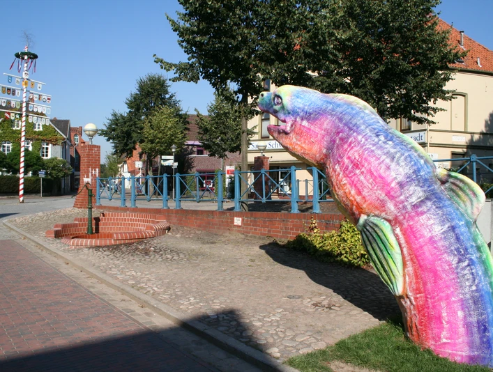 Bunte Skulptur eines Aals auf einem Gehweg vor traditioneller Backsteinarchitektur und blauem Himmel.