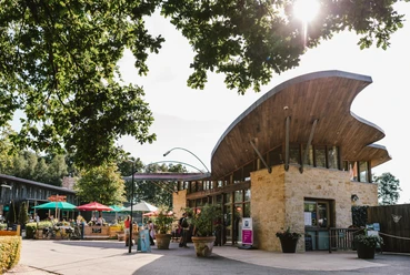 Besucherzentrum am Dreiländereck mit geschwungenem Holzdach, darunter Menschen bei sonnigem Wetter, umgeben von Natur und bunten Sonnenschirmen.