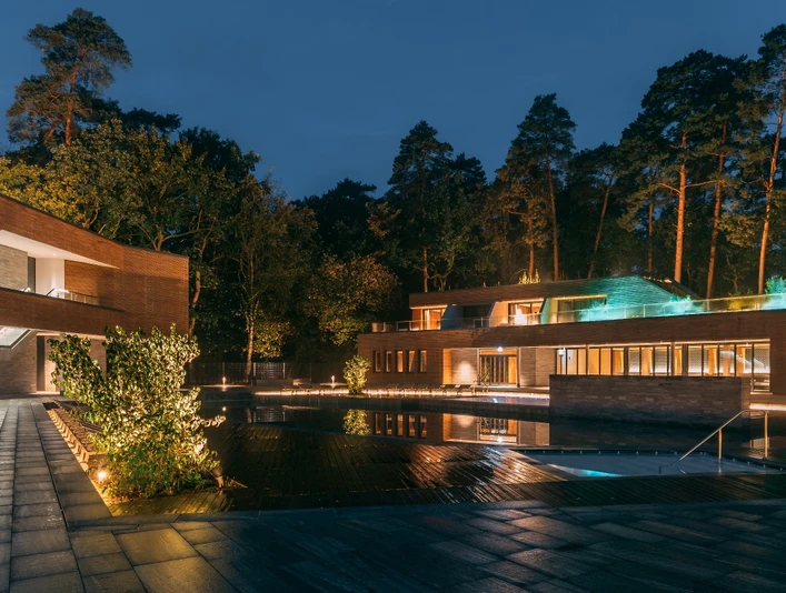 Eine moderne Wellnessanlage im Wald, nachts beleuchtet, umgeben von hohen Bäumen und einem Teich.