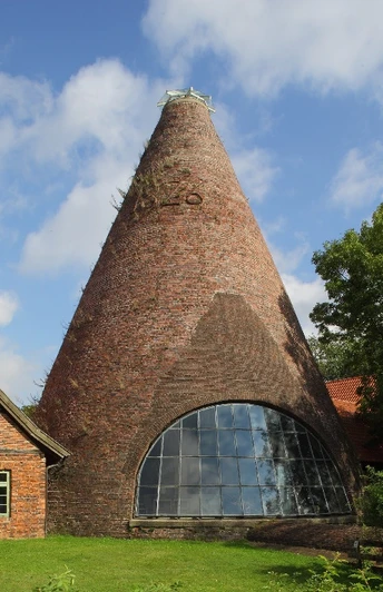 Historische Backstein-Glashütte mit großem konischen Schornstein, umgeben von Bäumen und Grünflächen.