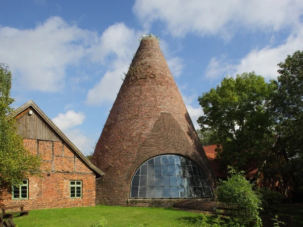 Historische Backstein-Glashütte mit großem konischen Schornstein, umgeben von Bäumen und Grünflächen.