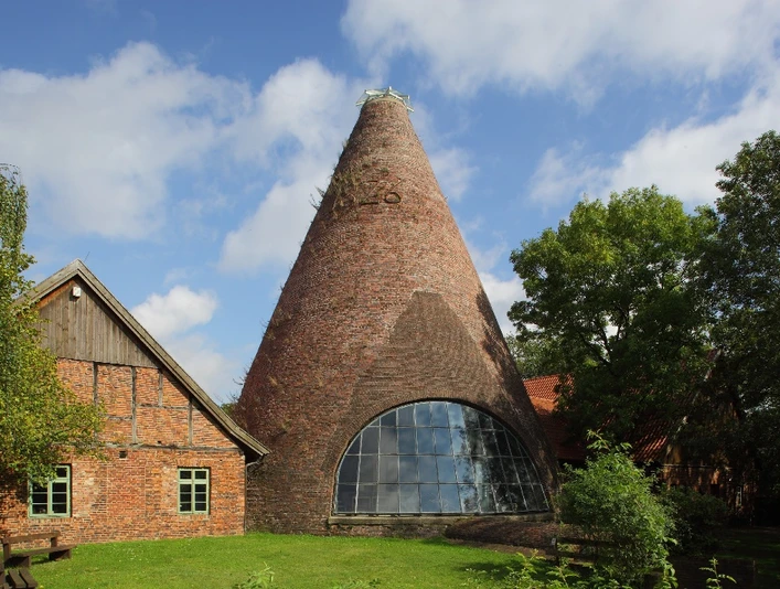 Historische Backstein-Glashütte mit großem konischen Schornstein, umgeben von Bäumen und Grünflächen.