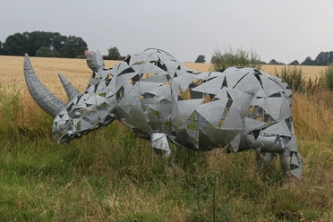 Eine moderne Nashorn-Skulptur aus Metallplatten steht in einer ländlichen Wiesenlandschaft.