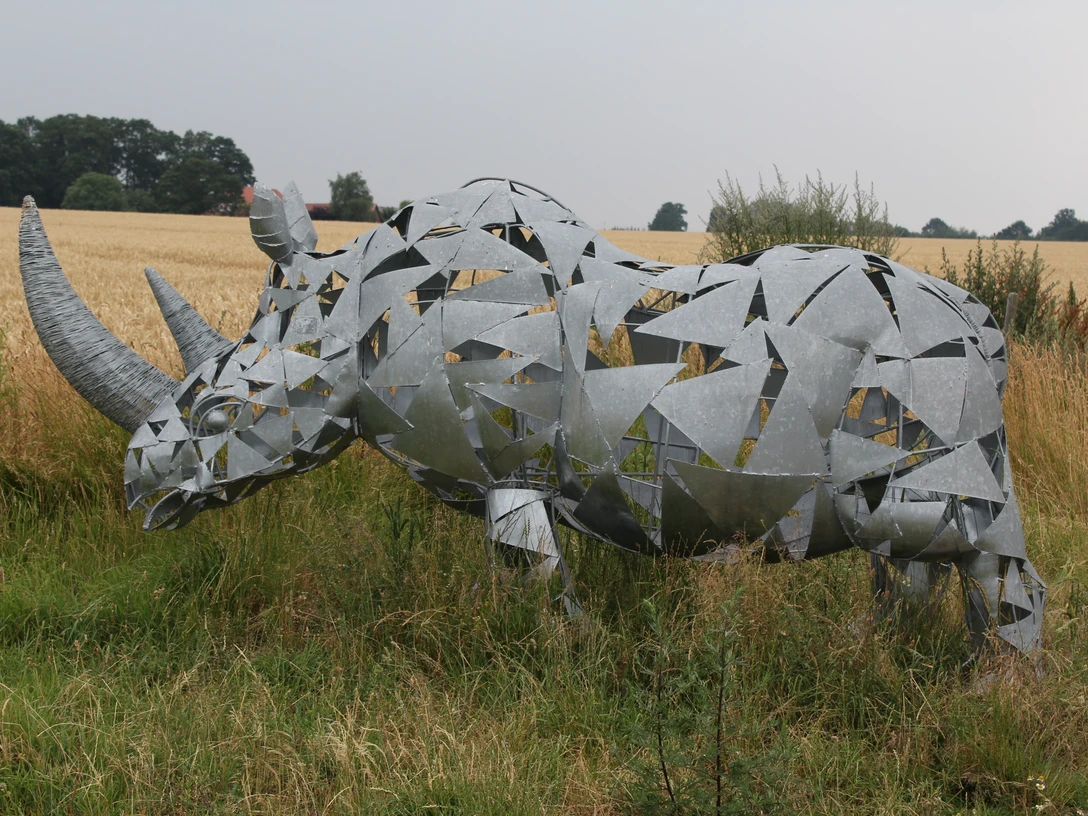 Eine moderne Nashorn-Skulptur aus Metallplatten steht in einer ländlichen Wiesenlandschaft.