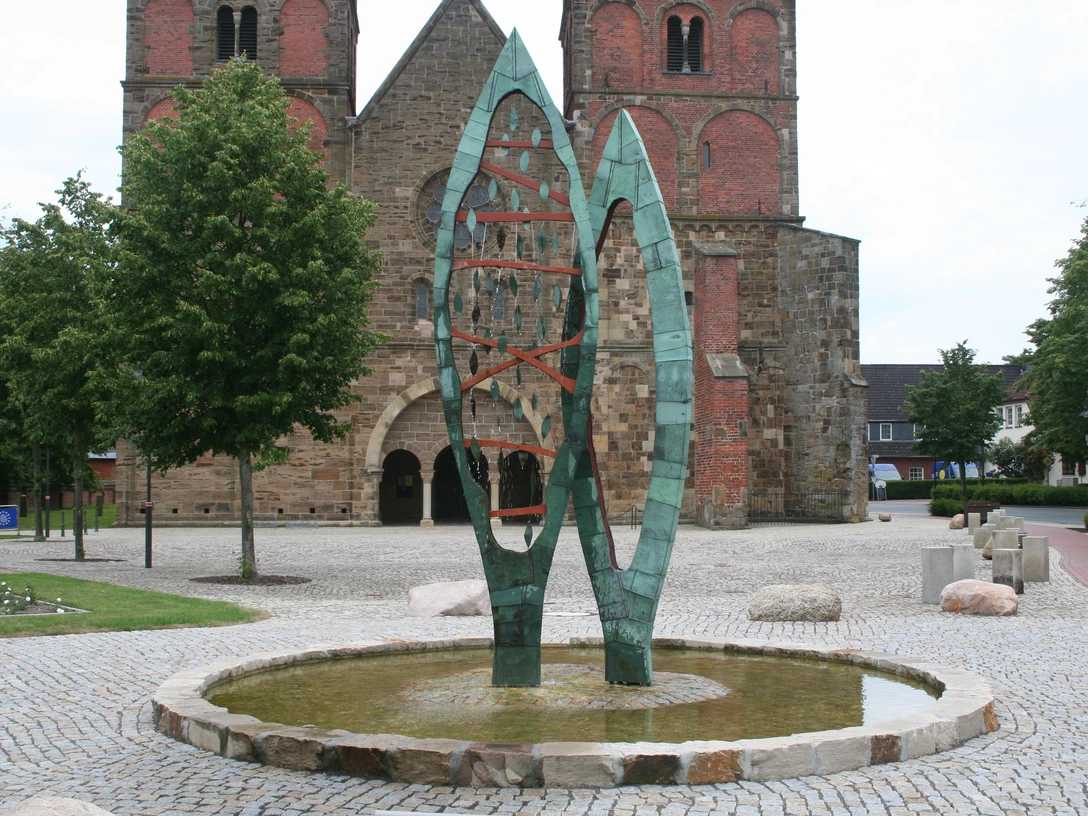 Skulptur aus grünem Metall, bestehend aus zwei geschwungenen Formen, auf einem gepflasterten Platz.