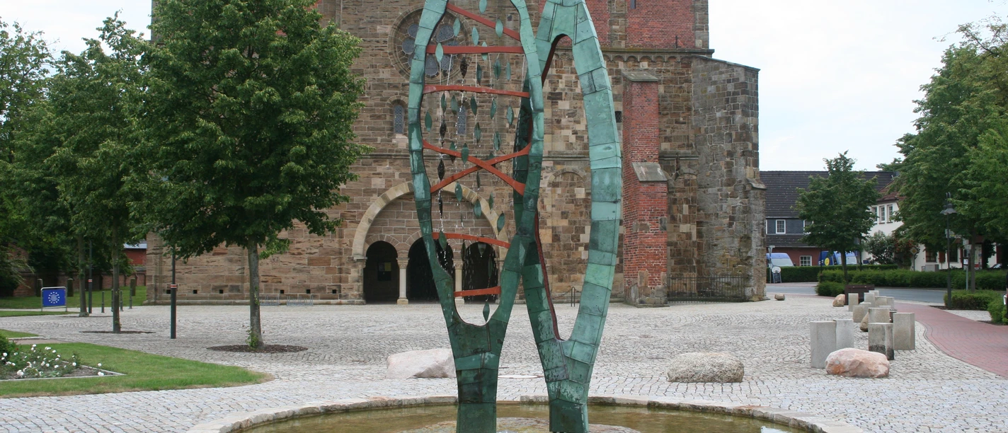 Skulptur aus grünem Metall, bestehend aus zwei geschwungenen Formen, auf einem gepflasterten Platz.