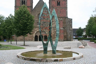 Brunnenskulptur STATIO Skulptur aus grünem Metall, bestehend aus zwei geschwungenen Formen, auf einem gepflasterten Platz.
