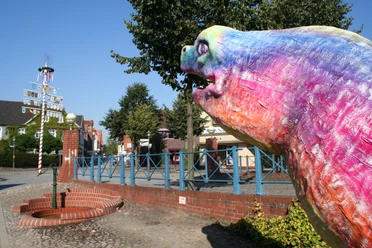 Bunte Aalskulptur in Hoya, Niedersachsen, mit Maibaum und Bäumen im Hintergrund, bei klarem Himmel.
