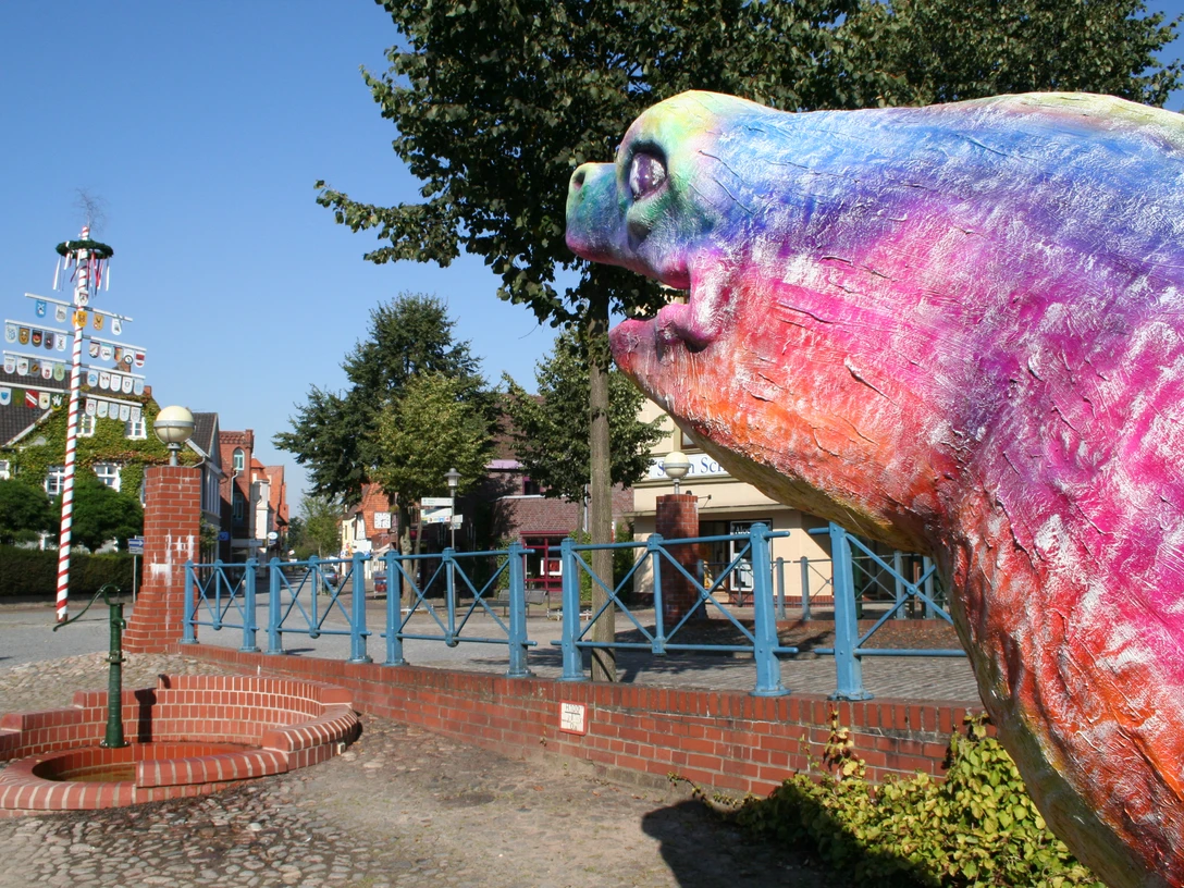 Bunte Aalskulptur in Hoya, Niedersachsen, mit Maibaum und Bäumen im Hintergrund, bei klarem Himmel.