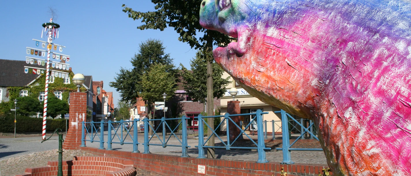 Bunte Aalskulptur in Hoya, Niedersachsen, mit Maibaum und Bäumen im Hintergrund, bei klarem Himmel.