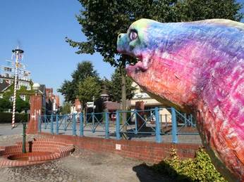 Bunte Aalskulptur in Hoya, Niedersachsen, mit Maibaum und Bäumen im Hintergrund, bei klarem Himmel.