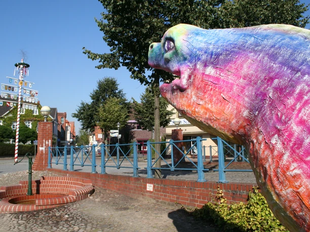Bunte Aalskulptur in Hoya, Niedersachsen, mit Maibaum und Bäumen im Hintergrund, bei klarem Himmel.