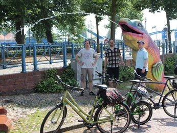 Drei Personen stehen neben gemeisterten Fahrrädern vor einer großen, farbenfrohen Fischskulptur.