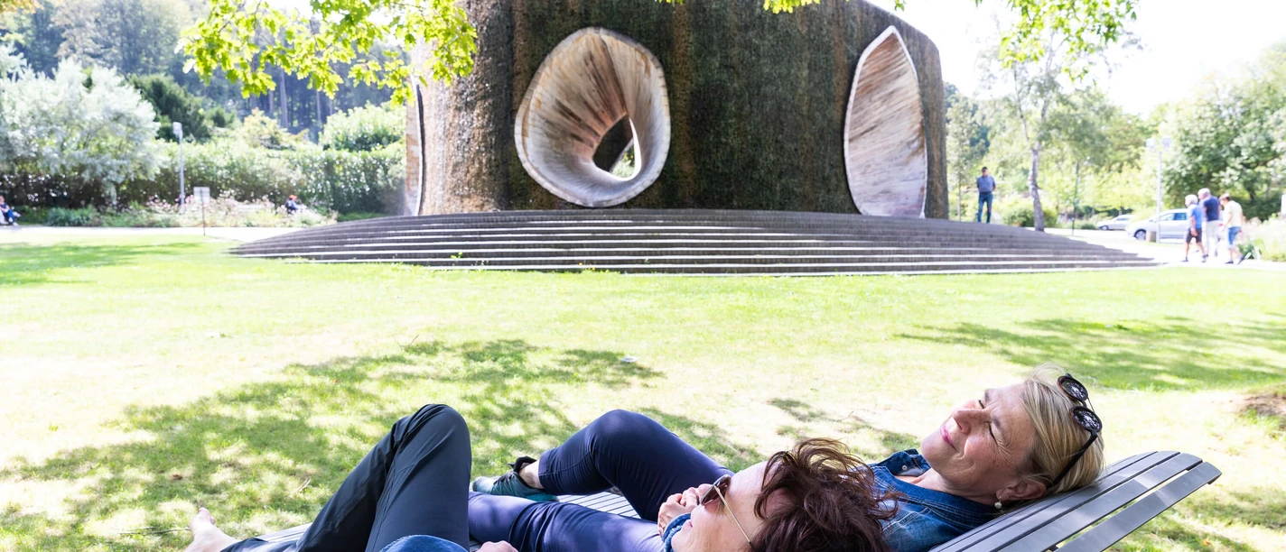 SoleArena Bad Essen Zwei Frauen entspannen auf einer Bank vor einem großen, skulpturalen Brunnen im Park.