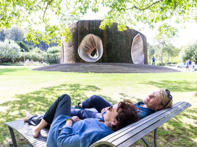 SoleArena Bad Essen Zwei Frauen entspannen auf einer Bank vor einem großen, skulpturalen Brunnen im Park.Two women relax on a bench in front of a large, sculptural fountain in the park.To kvinder slapper af på en bænk foran et stort, skulpturelt springvand i parken.Twee vrouwen relaxen op een bankje voor een grote, sculpturale fontein in het park.
