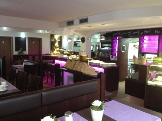 Cafe Pfeil Gemütlich eingerichtetes Café mit leuchtendem rosa Licht, dunkelbraunen Möbeln und gedeckten Tischen.