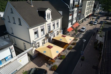 Haus Kranenberg Straßenansicht mit mehreren weißen Gebäuden, roten Markisen und gelben Sonnenschirmen vor Cafés.