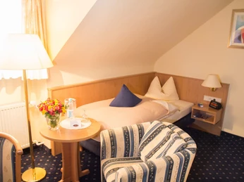 Zimmerbeispiel - Landhotel May-Hof Ein gemütliches Einzelzimmer mit Dachschräge, Bett, Sessel, Tisch mit Blumenstrauß und Lampe.