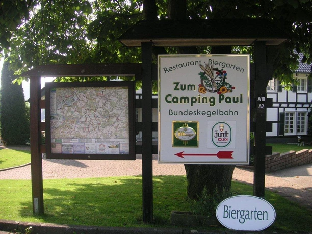 Hinweistafel Wandern Ein Holzschild unter Bäumen zeigt den Weg zum Restaurant und Biergarten "Zum Camping Paul" mit Kartenübersicht.