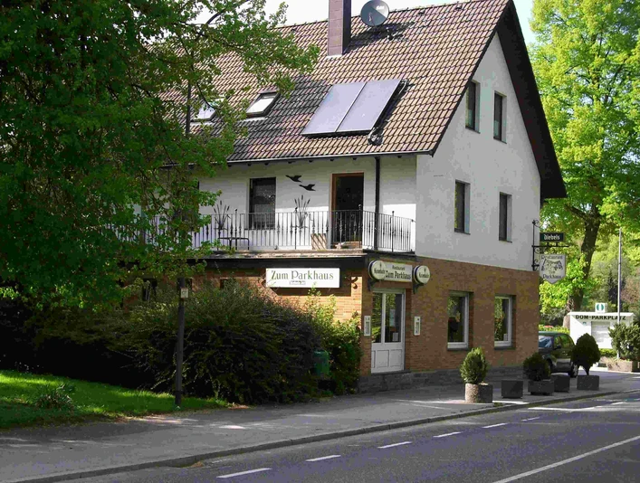 Restaurant "Zum Parkhaus" Gasthaus "Zum Parkhaus" mit Ziegeldach, weißen Wänden, umgeben von Bäumen und einer Straße davor.