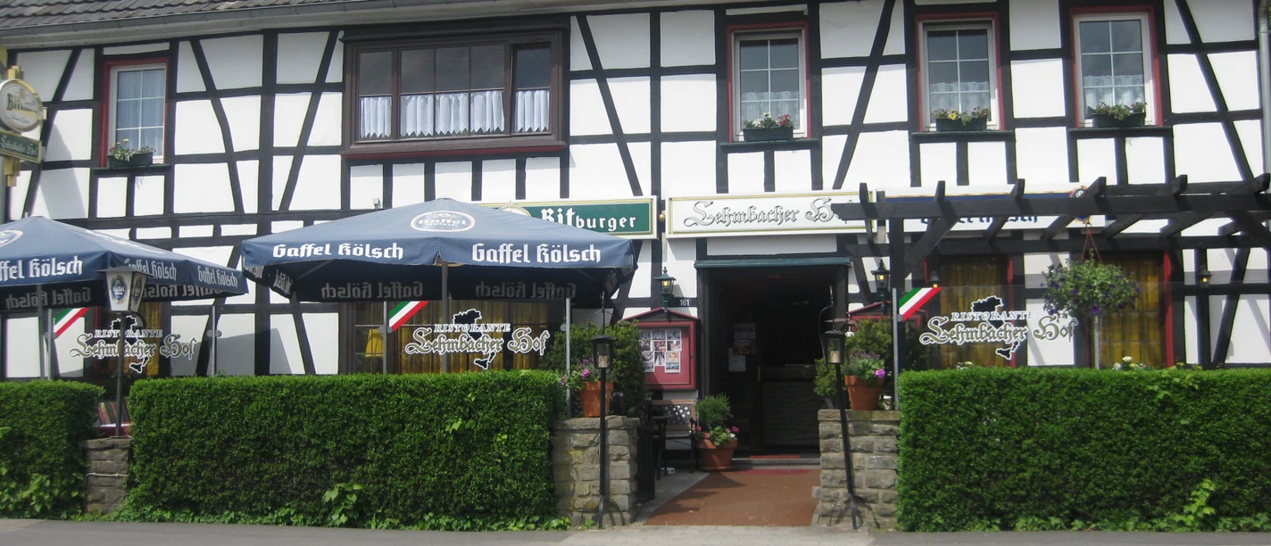 Lehmbacher Hof Fachwerkhaus mit Restaurant. Vor dem Gebäude stehen Biergarten-Sonnenschirme und grüne Hecke.