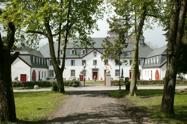 Außenansicht Herrenhaus mit weißen Fassaden und roten Türen, flankiert von Bäumen und gepflegter Rasenfläche.