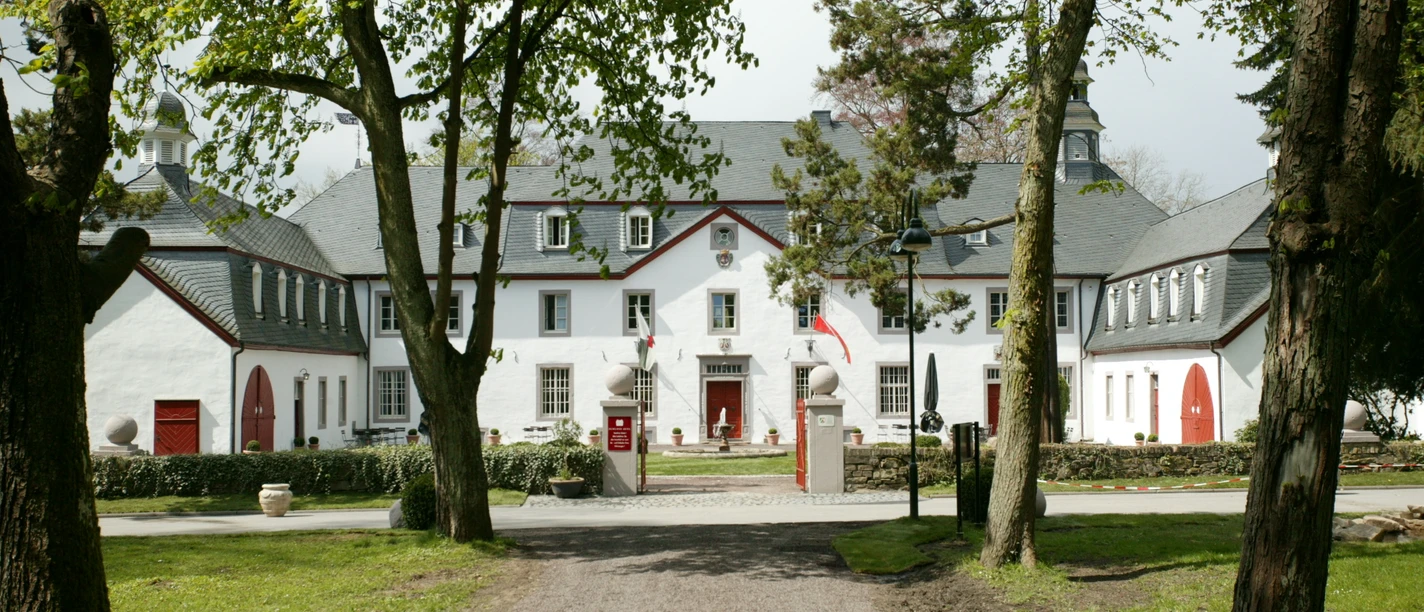 Außenansicht Herrenhaus mit weißen Fassaden und roten Türen, flankiert von Bäumen und gepflegter Rasenfläche.