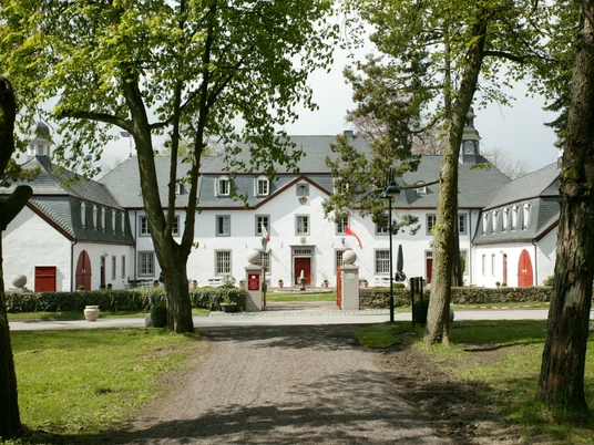 Außenansicht Herrenhaus mit weißen Fassaden und roten Türen, flankiert von Bäumen und gepflegter Rasenfläche.