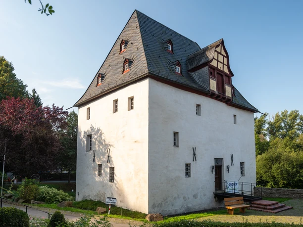 Golfplatz Burg Overbach <p>Historisches Fachwerkhaus im Sonnenschein, umgeben von grüner Vegetation und blühenden Bäumen.</p>