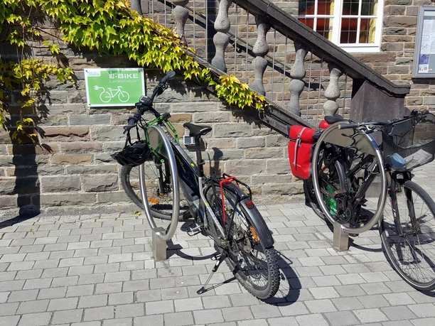 Ladestation am Rathaus Zwei Fahrräder, eins mit Packtaschen, parken an einer Steinmauer, vor einer Treppe und einem E-Bike-Schild.