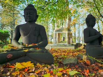 Buddhastatuen im Garten Zwei sitzende Buddha-Statuen im herbstlichen Park, umgeben von buntem Laub und Bäumen.