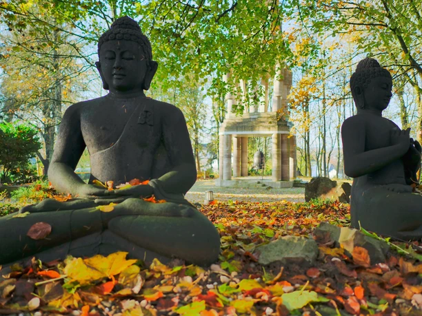 Buddhastatuen im Garten Zwei sitzende Buddha-Statuen im herbstlichen Park, umgeben von buntem Laub und Bäumen.