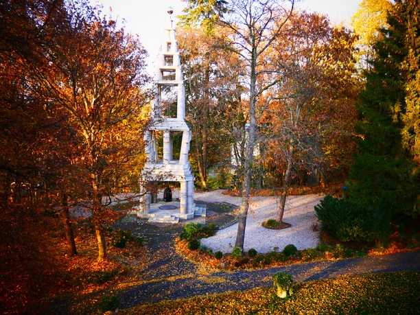 Garten im Herbst Ein steinerner Glockenturm im farbenfrohen Herbstwald, umgeben von Laub und Spazierwegen.