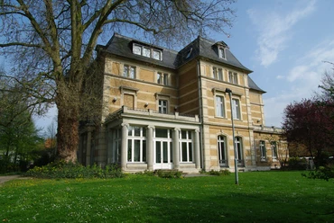 Villa Zanders Historische Villa mit gelber Fassade, großen Fenstern und Mansardendach, umgeben von Bäumen und Rasen.