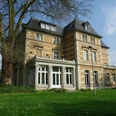 Villa Zanders Historische Villa mit gelber Fassade, großen Fenstern und Mansardendach, umgeben von Bäumen und Rasen.