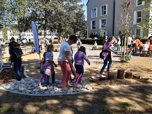Barfußpfad Eine Gruppe von Kindern und Erwachsenen nutzt einen Spielplatz mit Holzstämmen und Kieselsteinen.