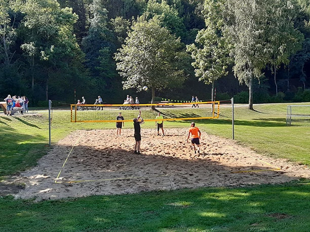 Volleyballfeld Vier Personen spielen Beachvolleyball auf einem Sandplatz in einem grünen Park bei sonnigem Wetter.