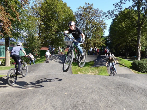 Pumptrack in Morsbach Mehrere Radfahrer auf BMX-Bikes fahren und springen in einem belebten Park bei sonnigem Wetter.