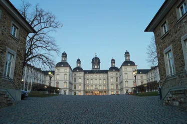 Portal Schloss Bensberg <p>Das Bild zeigt das Schloss Bensberg bei Dämmerung, mit einem gepflasterten Vorplatz aus der Frontalansicht.</p>