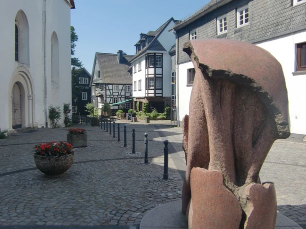 Station an der Kirche im Ortszentrum Nümbrecht Das Bild zeigt eine malerische Altstadtgasse in Nümbrecht mit Skulptur im Vordergrund.
