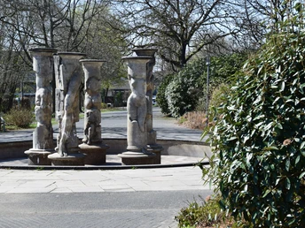 Kurpark in Nümbrecht  Skulpturengruppe aus Steinsäulen mit Reliefs auf einem Platz, umgeben von Bäumen und Sträuchern.