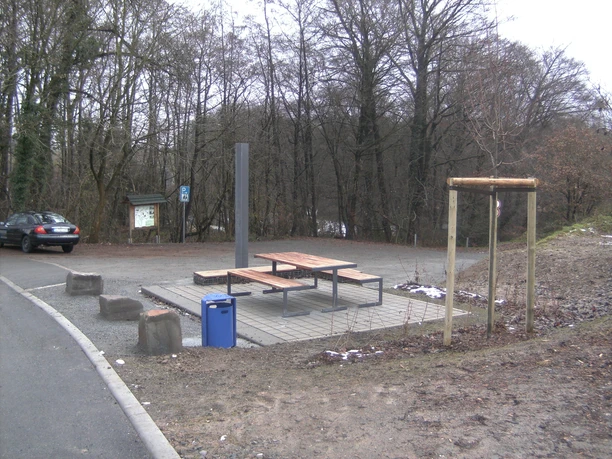 DIGITAL CAMERA Ein ruhiger Rastplatz im Wald mit einem Holztisch, Bänken und einer blauen Mülltonne im Vordergrund.