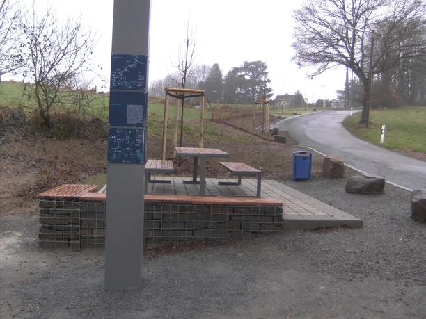 DIGITAL CAMERA Picknickplatz mit Tisch und Bänken auf gepflastertem Untergrund, umgeben von kahlen Bäumen und Wiese.