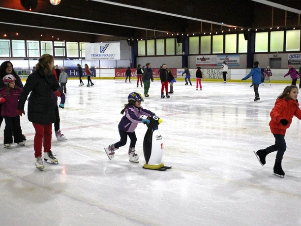 Eissporthalle Wiehl Kinder und Erwachsene beim Eislaufen in einer belebten Eishalle mit Unterstützungspinguinen.