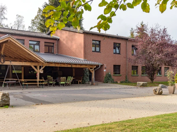 Gut Ommeroth Backstein-Gebäude mit überdachter Terrasse, Sitzgelegenheiten und umgebender Natur im Vordergrund.