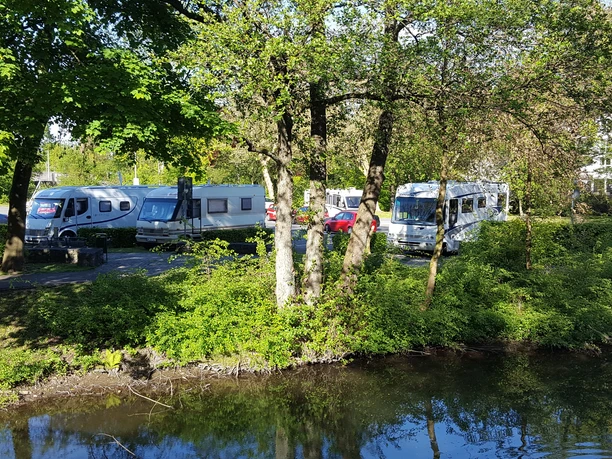 Wohnmobilstellplatz Ohler Wiesen Wohnmobile parken unter Bäumen auf einem Campingplatz, umgeben von viel Grün und einem Gewässer.